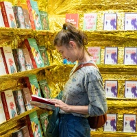 Frau liest gespannt in einem Buch vor einem goldenen Bücherregal auf der Buchmesse 2025 Frau liest gespannt in einem Buch vor einem goldenen Bücherregal auf der Buchmesse 2025
