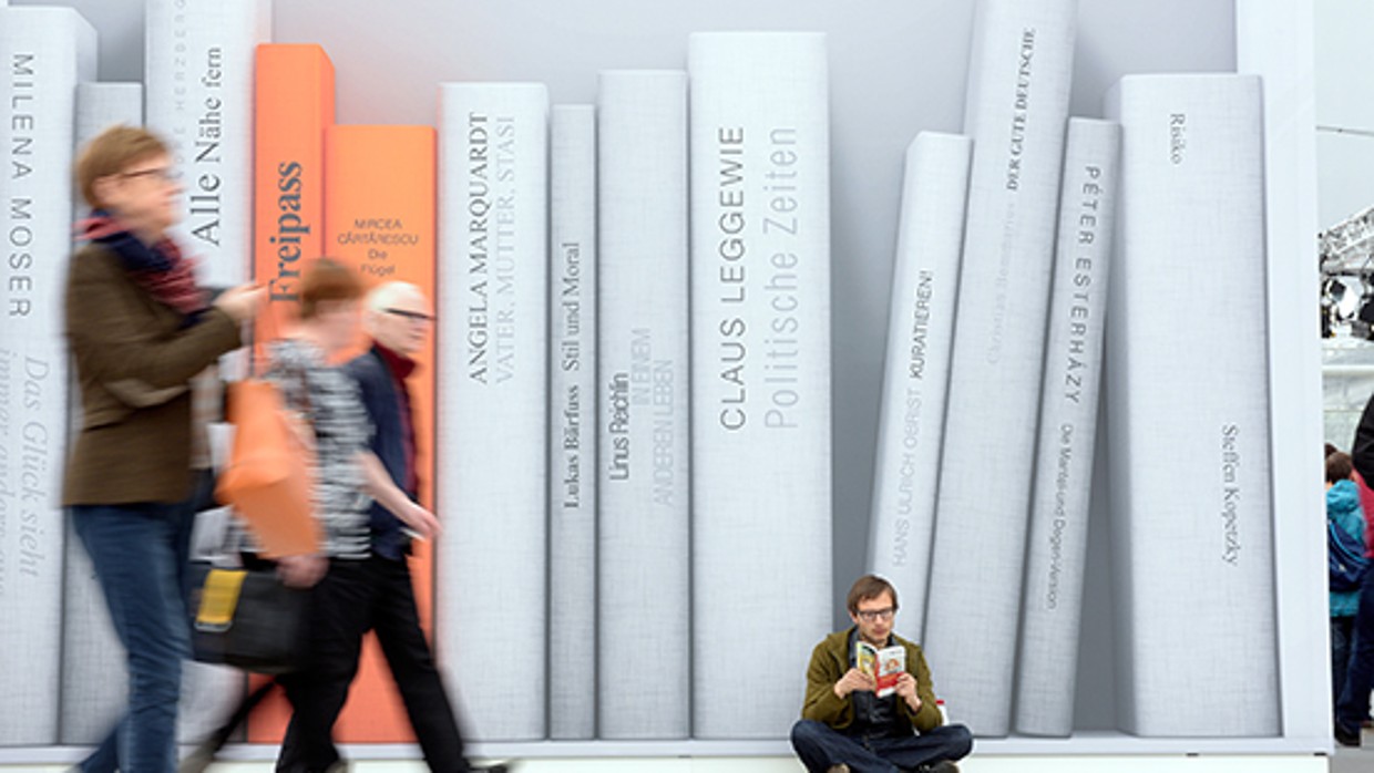 Ein Besucher sitzt im Schneidersitz vor einer Wand und liest in einem Buch. Auf der Wand im Hintergrund sind verschieden überdimensionale Bücher in schwarz weiß abgebildet. Zwei der Bücher sind Orange. 