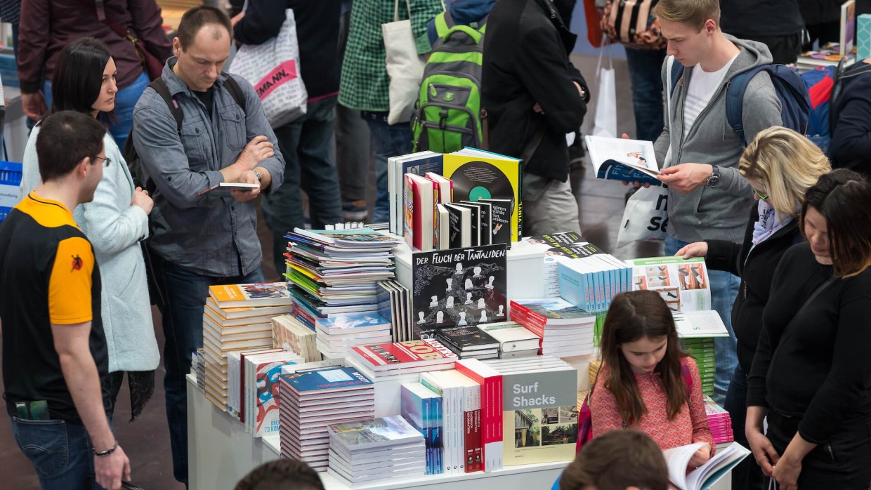 Leipziger Buchmesse 2017: Besucher:innen stehen um Bücherinsel herum und blättern in Büchern. Die Insel steht mittig im Bild. 