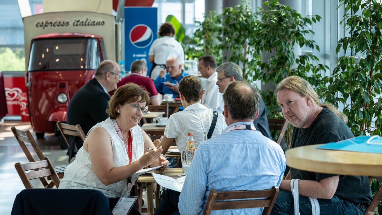 Besucher:innen sitzen um Tische herum. Im Hintergrund sieht man eine Person essen. Dahinter steht eine Kaffeebar.