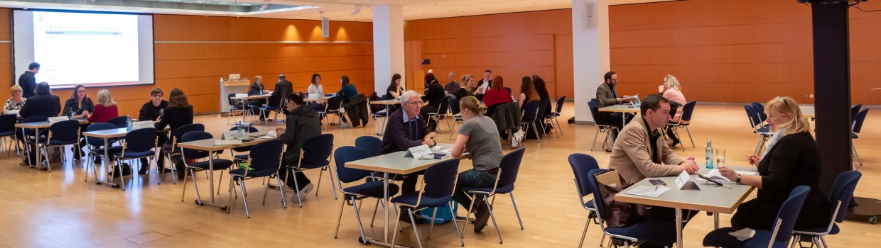 Eine Veranstaltung auf der Leipziger Buchmesse im Congress Center Leipzig. Im Raum stehen mehrere Tische an denen verschieden viele Personen sitzen. 