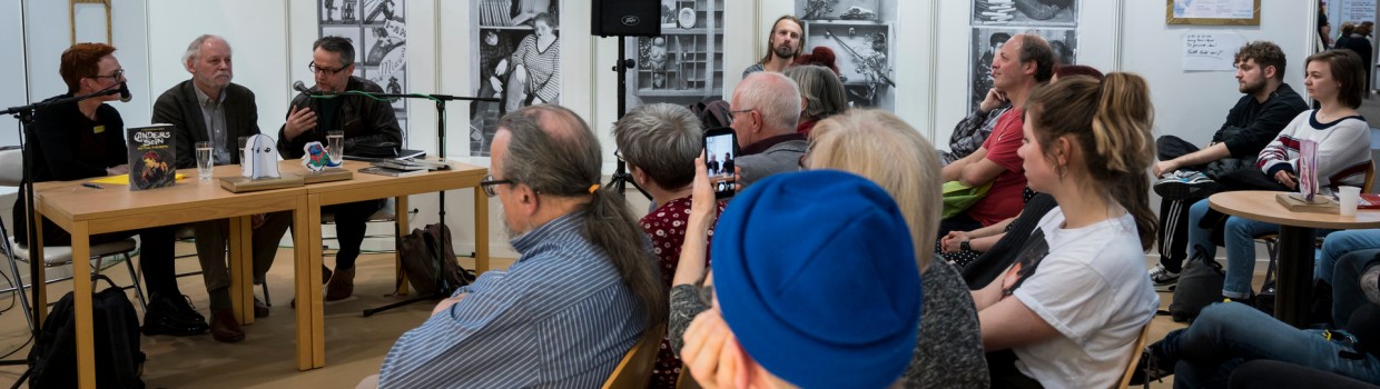 Eine Veranstaltung auf der Leipziger Buchmesse. Drei Personen sitzen an einem Tisch mit Mikrophon im Hintergrund. Im Vordergrund und im rechten Bild sitzen Besucher:innen. Eine Veranstaltung auf der Leipziger Buchmesse. Drei Personen sitzen an einem Tisch mit Mikrophon im Hintergrund. Im Vordergrund und im rechten Bild sitzen Besucher:innen.