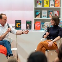In der #buchbar sitzt Marc Elsberg am Tresen und unterhält sich mit einer Frau. Dabei sitzen im Vordergrund Personen an Tischen und hören zu. Im Hintergrund sieht man Tresen und Regale, gefüllt mit Büchern. In der #buchbar sitzt Marc Elsberg am Tresen und unterhält sich mit einer Frau. Dabei sitzen im Vordergrund Personen an Tischen und hören zu. Im Hintergrund sieht man Tresen und Regale, gefüllt mit Büchern.