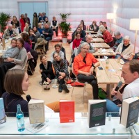 In der #buchbar sitzt im Vordergrund Marc Elsberg am Tresen und unterhält sich mit einer Frau. Dabei sitzen im Hintergrund viele verschiedene Personen an Tischen und auf dem Boden und Hören aufmerksam zu.  In der #buchbar sitzt im Vordergrund Marc Elsberg am Tresen und unterhält sich mit einer Frau. Dabei sitzen im Hintergrund viele verschiedene Personen an Tischen und auf dem Boden und Hören aufmerksam zu.
