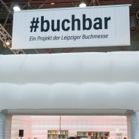 Im Eingang der #buchbar stehen mehrere Besucher, im inneren sitzen sie an den Tischen und lauschen Marc Elsberg, welcher am Tresen sitzt. Über dem Eingang hängt ein Banner mit #buchbar "Ein Projekt der Leipziger Buchmesse".  Im Eingang der #buchbar stehen mehrere Besucher, im inneren sitzen sie an den Tischen und lauschen Marc Elsberg, welcher am Tresen sitzt. Über dem Eingang hängt ein Banner mit #buchbar "Ein Projekt der Leipziger Buchmesse".