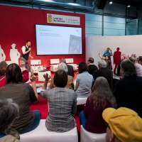 Eine Veranstaltung auf der Buchmesse. Ein Mann auf der Bühne spricht ins Mikrofon, ihm gegenüber sitzen Zuschauer:innen und heben teilweise die Hand. 