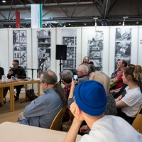 Eine Veranstaltung auf der Leipziger Buchmesse. Drei Personen sitzen an einem Tisch mit Mikrophon im Hintergrund. Im Vordergrund und im rechten Bild sitzen Besucher:innen. 