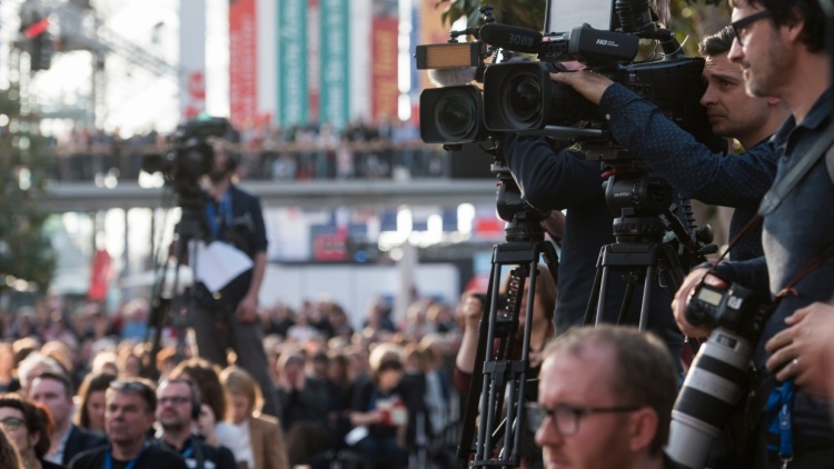 Ein Foto von einer Veranstaltung auf der Leipziger Buchmesse. Zu sehen sind drei Kameramänner und deren Kameras, welche auf ein Objekt außerhalb des Bildes gerichtet sind. Zwei der Männder sind scharf rechts im Bild zu sehen. Einer ist im Hintergrund verschwommen abgebildet. 