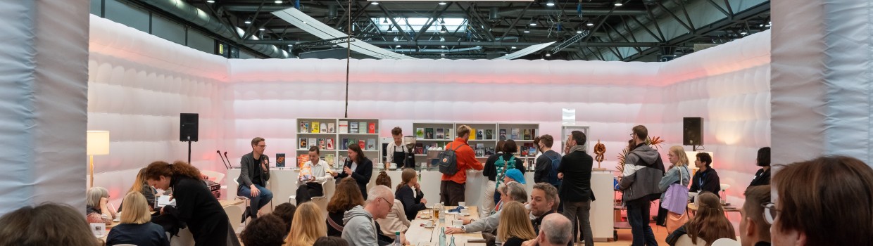 In der #buchbar sitzt das Publikum an langen Tischen während im Hintergrund Menschen an einer Bartheke zu sehen sind. An der gleichen Bar sitzen links 3 Personen mit Mikrophonen im Gespräch.