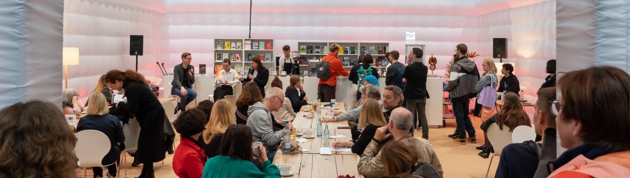 In der #buchbar sitzt das Publikum an langen Tischen während im Hintergrund Menschen an einer Bartheke zu sehen sind. An der gleichen Bar sitzen links 3 Personen mit Mikrophonen im Gespräch.