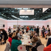 In der #buchbar sitzt das Publikum an langen Tischen während im Hintergrund Menschen an einer Bartheke zu sehen sind. An der gleichen Bar sitzen links 3 Personen mit Mikrophonen im Gespräch. In der #buchbar sitzt das Publikum an langen Tischen während im Hintergrund Menschen an einer Bartheke zu sehen sind. An der gleichen Bar sitzen links 3 Personen mit Mikrophonen im Gespräch.
