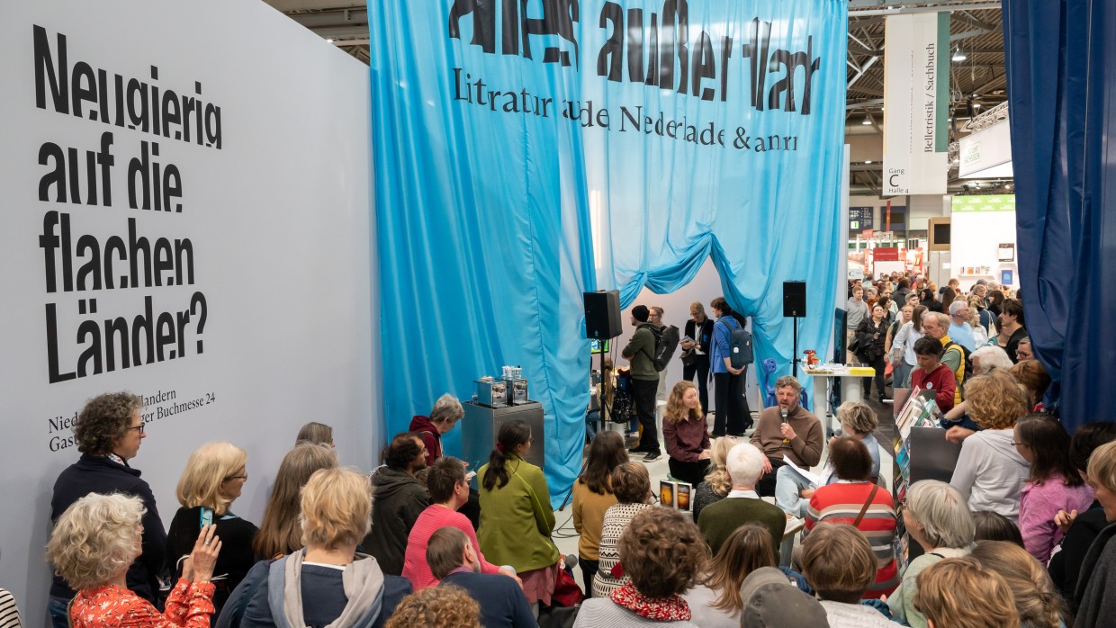 Der Stand des Gastlandes 2024 Niederlande und Flandern. Im Himtergrund hängt ein Blauer Vorhang mit der Aufschrift "alles außer flach". Im Vordergrund hören viele Menschen der Lesung zu.