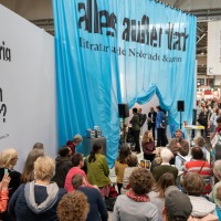 Der Stand des Gastlandes 2024 Niederlande und Flandern. Im Himtergrund hängt ein Blauer Vorhang mit der Aufschrift "alles außer flach". Im Vordergrund hören viele Menschen der Lesung zu.