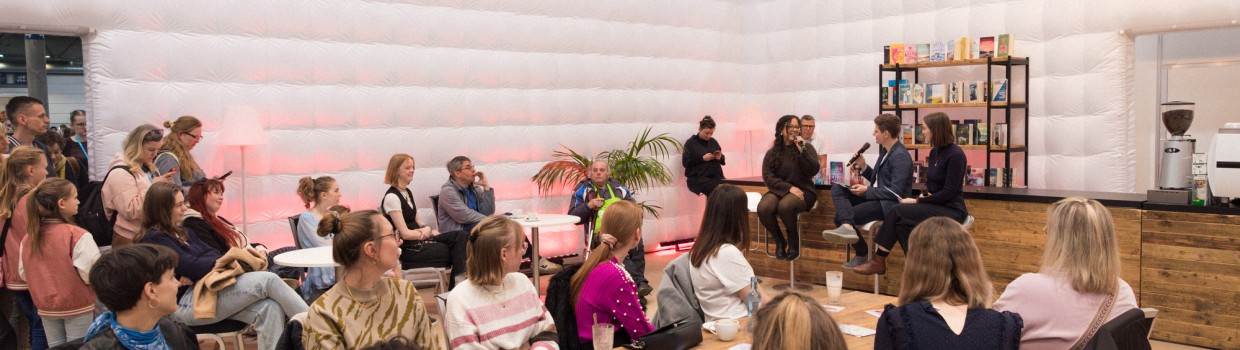 Eine Autorin im Gespräch mit den beiden Moderator:innen in der #buchbar während der Leipziger Buchmesse 2025 vor vollbesetztem Publikum