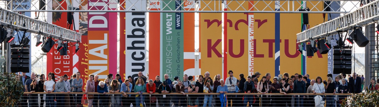 viele Menschen stehen auf der Brücke in der Glashalle der Messe hinter ihnen hängen viele Flaggen