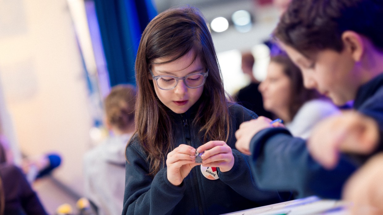 mehrere Kinder sitzen an einem Tisch, ein Mädchen betrachtet interessiert ein Objekt in ihren Händen während der Leipziger Buchmesse 2025