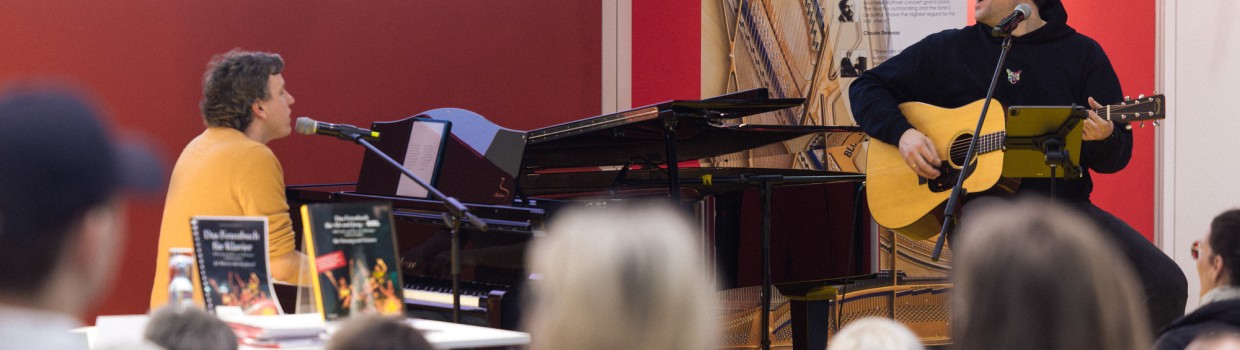 Das Musik-Café der Leipziger Buchmesse 2025 mit einer Bühnenveranstaltung von zwei Musikern an Gitarre und Klavierflügel vor vollbesetztem Publikum