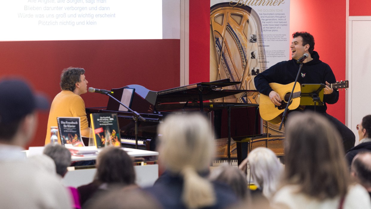Das Musik-Café der Leipziger Buchmesse 2025 mit einer Bühnenveranstaltung von zwei Musikern an Gitarre und Klavierflügel vor vollbesetztem Publikum