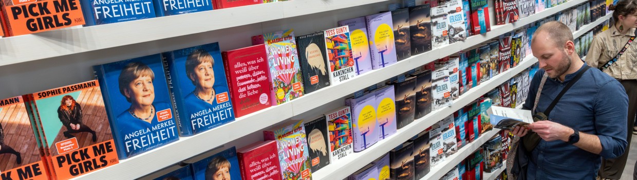 buntes Bücherregal mit vielen Büchern, vor dem Besucher:innen der Leipziger Buchmesse 2025 stehen und lesen
