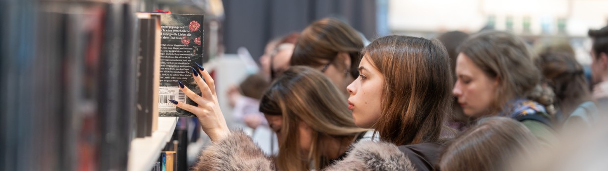 junge Frau entnimmt ein Buch aus einem gefüllten Buchregal auf der Leipziger Buchmesse 2026