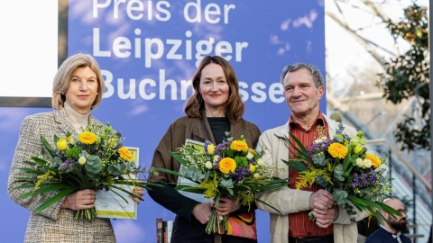 Die drei Preisträger:innen des Preises der Leipziger Buchmesse 2026 stehen gemeinsam mit Blumensträußen und Urkunden in den Händen auf der Bühne