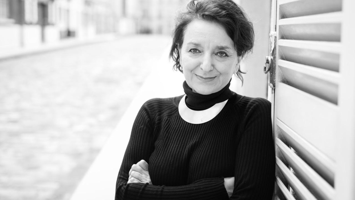 Ein schwarz-weiß Portrait von Eva Illouz. Sie steht mit verschränkten Armen an eine Wand gelehnt im Freien und lächelt in die Kamera.