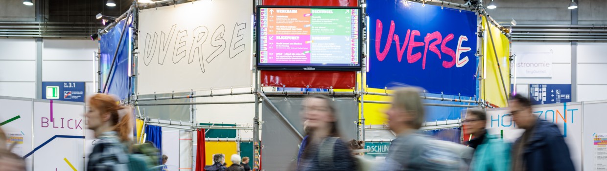 Foto vom UVERSE Stand der LBM23 mit Besucher:innen, die im Vordergrund daran vorbei laufen