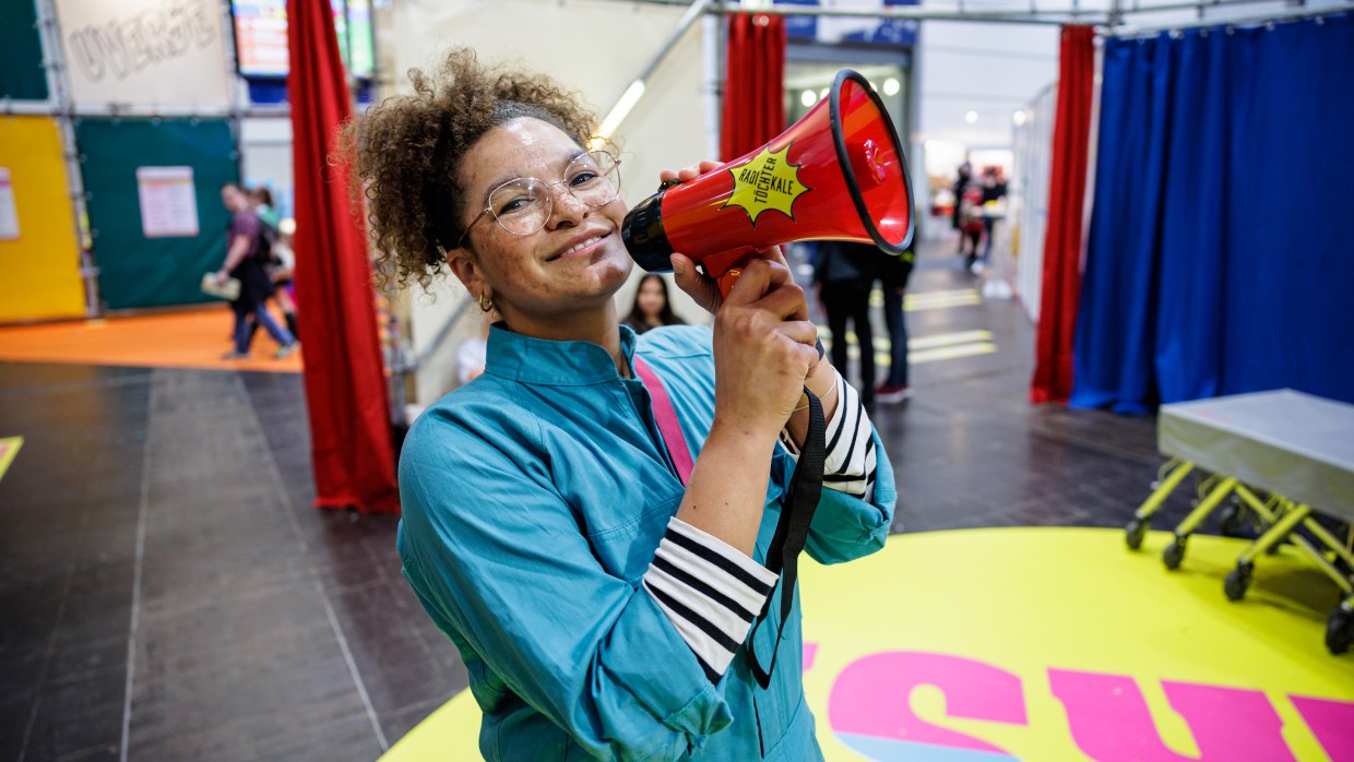 Foto aus dem UVERSE Programm der LBM23 von einer bunt gekleideten Frau, die ein rotes Megaphon hält