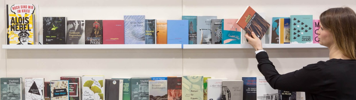 A woman is standing in front of a bookshelf and reaching for a book. The woman is dressed in black and is standing on the right side of the picture. The bookshelf consists of four white shelves. 