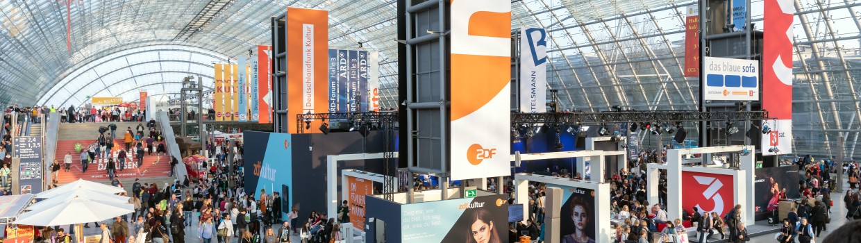 The glass hall from the inside with many visitors and stands. In the middle, there are many media banners hanging.
