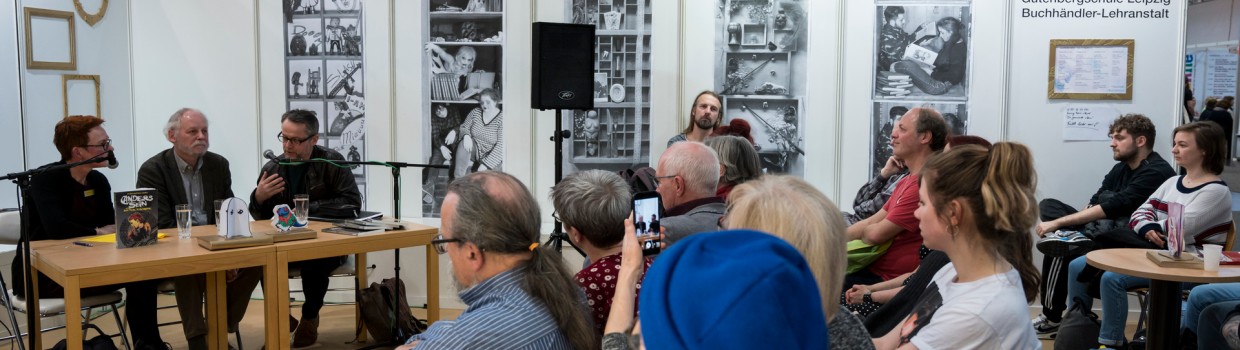 An event at the Leipzig Book Fair. Three people are seated at a table with a microphone in the background. Visitors are seated in the foreground and on the right side of the picture