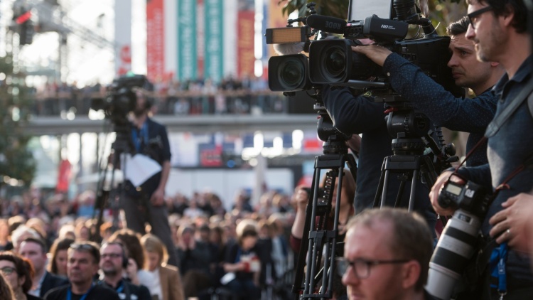 A photo from an event at the Leipzig Book Fair. Three cameramen and their cameras can be seen pointing at an object outside the frame. Two of the men are clearly visible on the right side of the image. One is blurred in the background. 