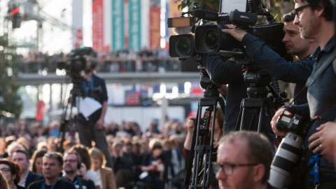 A photo from an event at the Leipzig Book Fair. Three cameramen and their cameras can be seen pointing at an object outside the frame. Two of the men are clearly visible on the right side of the image. One is blurred in the background. 