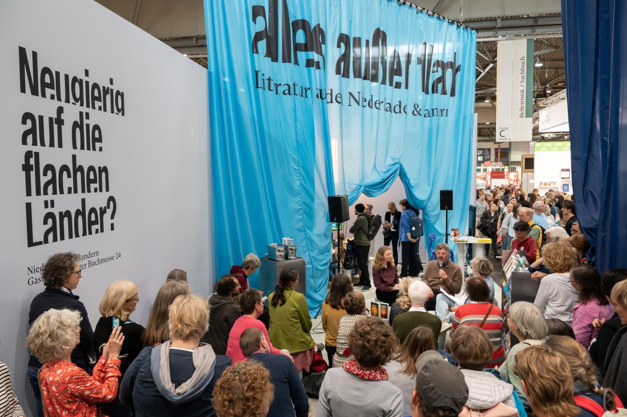 The stand of the host country for 2024, the Netherlands and Flanders. In the background hangs a blue curtain with the inscription “alles außer flach”. In the foreground, many people listen to the reading.