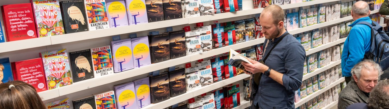 A man stands reading a book in front of a large bookcase at the Leipzig Book Fair in 2025