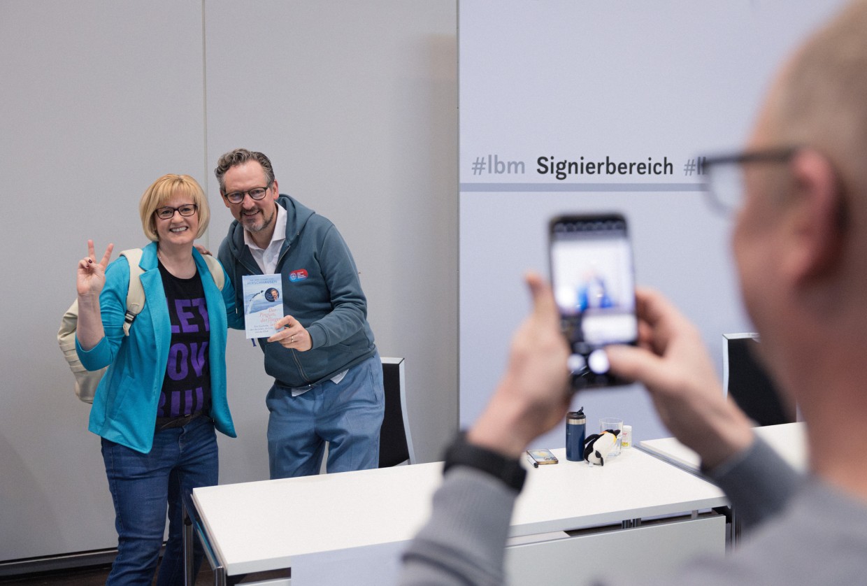 A woman stands next to an author in the signing area at the Leipzig Book Fair. Both laugh at the camera. 