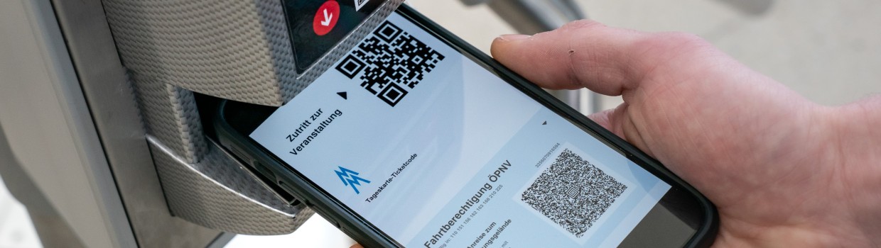 One hand holds a smartphone under the scanner at the turnstile entrance. The display shows an electronic ticket for the Leipzig Trade Fair with a QR code, including a public transport ticket.
