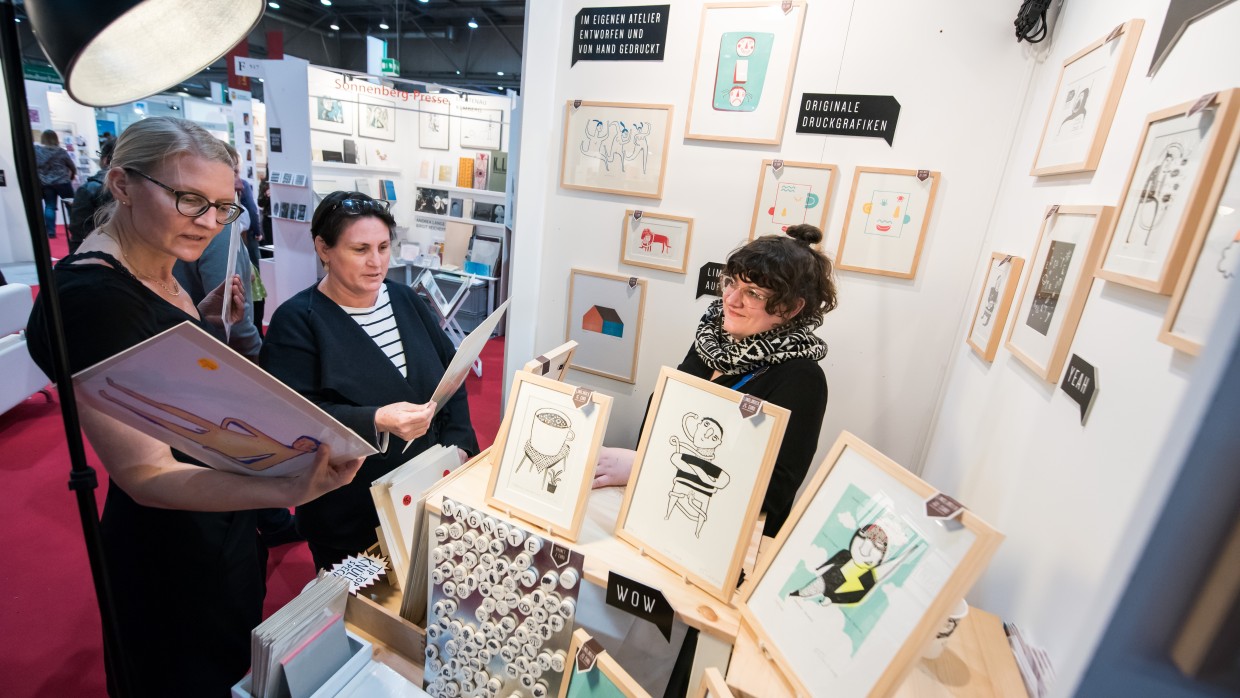 Zwei Besucherinnen schauen sich Grafiken an einem Messestand an. Im Messestand sitzt eine Frau, um Sie herum sind viele verschiedene Grafiken und Kunstdrucke zu sehen. 